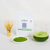 HINOKA Pure matcha powder with a whisk, bowl of matcha powder, and glass of green tea on a white background