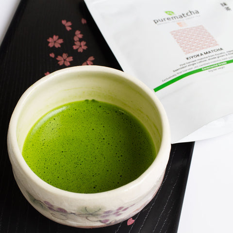Ceramic cup of green matcha tea with a package labeled 'purematcha' in the background.