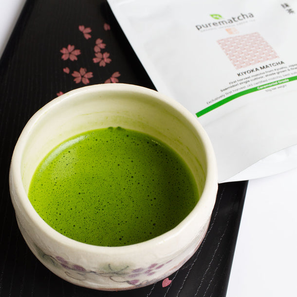 Ceramic cup of green matcha tea with a package labeled 'purematcha' in the background.