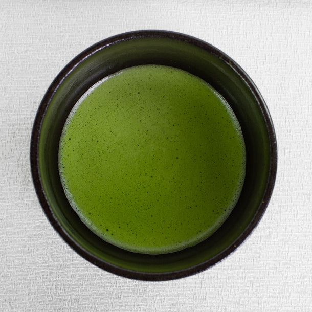 Pure Matcha ceremonial grade whisking in a dark ceramic bowl on a light fabric background