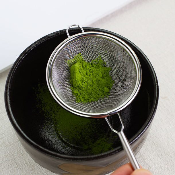 Ceremonial grade Japanese Matcha Tea powder being sifted into a black bowl using a metal sieve.