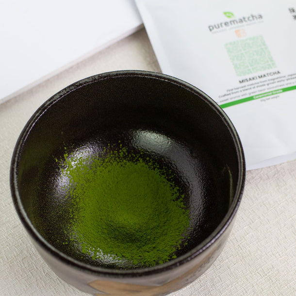Black ceramic cup filled with green matcha powder next to a package labeled 'purematcha'.