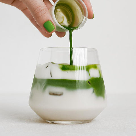 Hand pouring green liquid  from matcha mini shaker bottle into a glass with ice on a white background